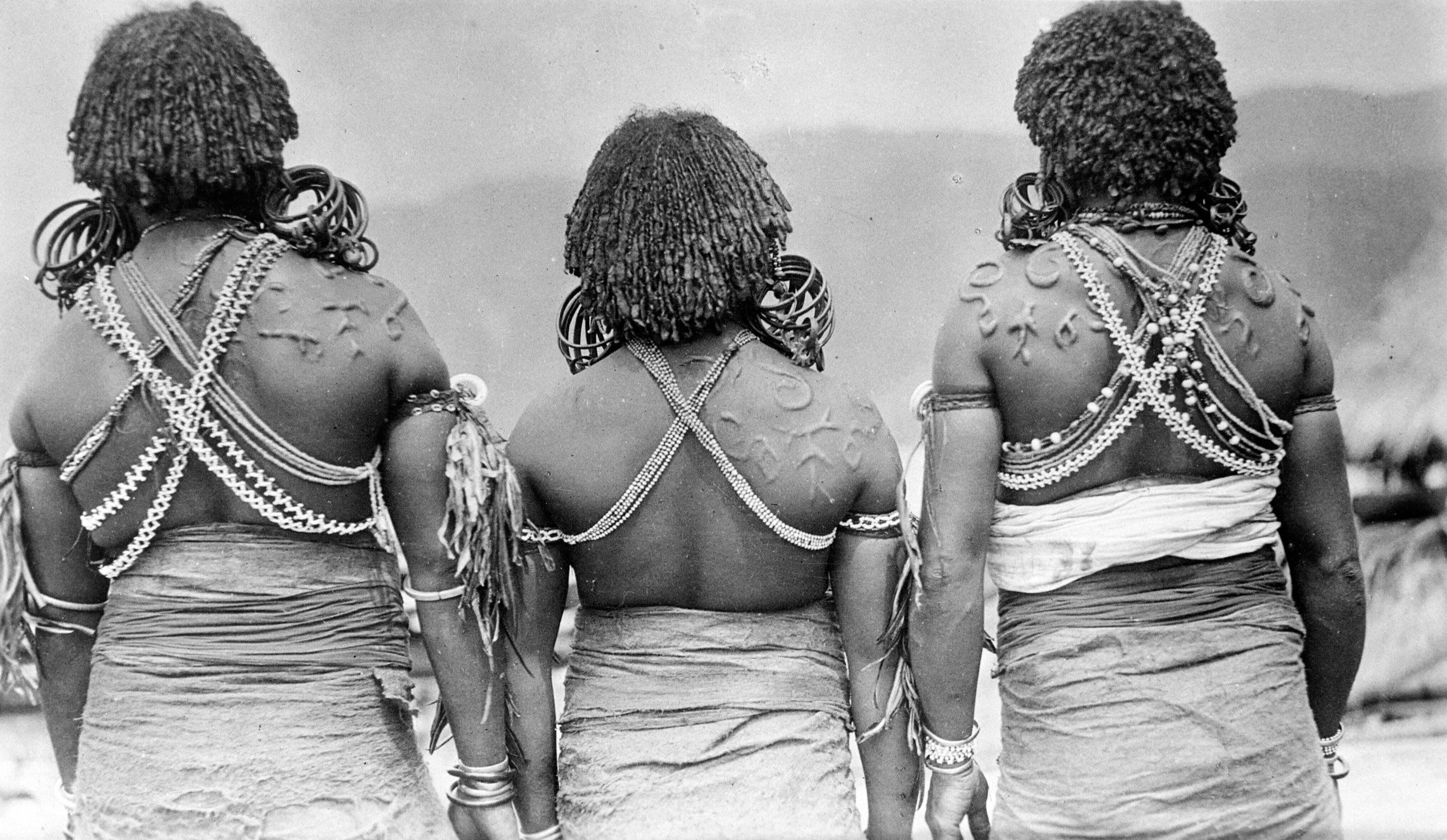 Portrait of three Papuan women of the Sentani Lake region with scarifications and arm, ear, neck and back ornaments, photographed from the back